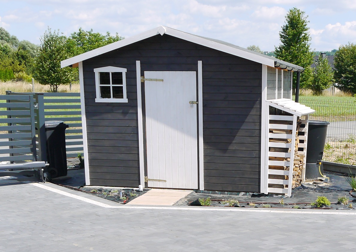Mała drewniana konstrukcja w kolorze grafitowym z białymi detalami, w tym drzwi i okno, z dobudowanym składem na drewno, umieszczona na kostce brukowej z widocznym ogrodem i drzewami w tle.