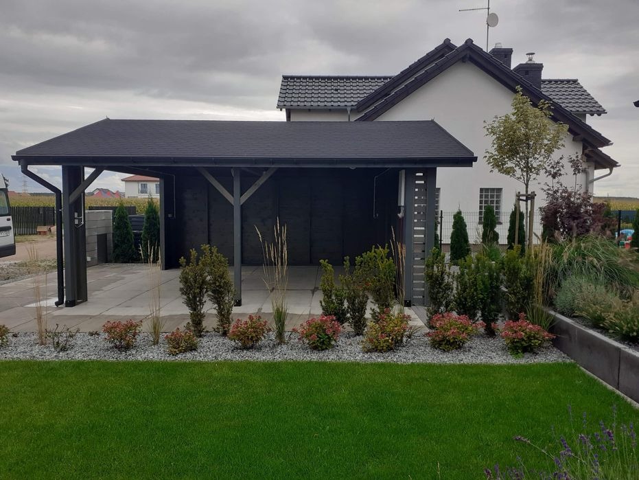 Zadaszone miejsce parkingowe z ciemnego drewna, z płaskim dachem, przylegające do domu z białymi ścianami, ozdobione rabatami z krzewów i traw.