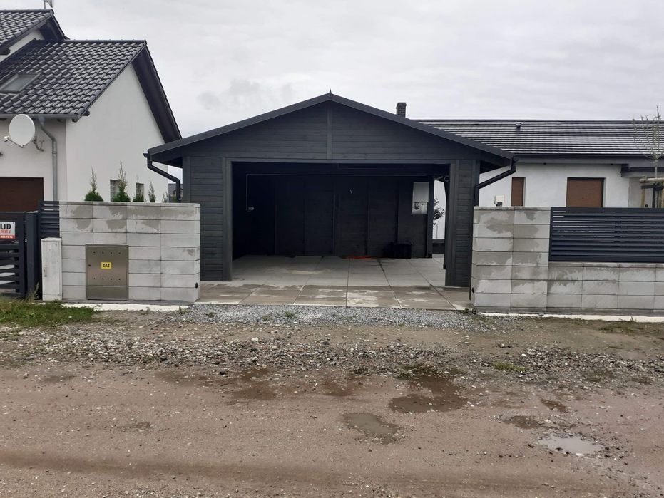 Nowoczesna, ciemnoszara wiata garażowa z płaskim dachem, wykończona drewnianą elewacją, na tle dwóch domów jednorodzinnych i betonowego ogrodzenia.