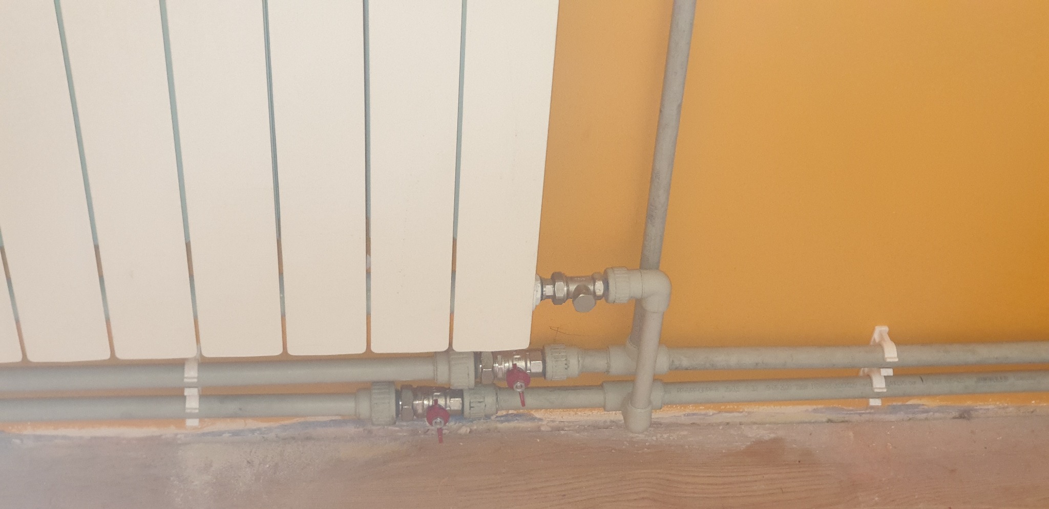 Biały grzejnik panelowy podłączony do instalacji z rur plastikowych z zaworami z czerwonymi pokrętłami, widoczne mocowania rur do ściany w pomarańczowym kolorze.