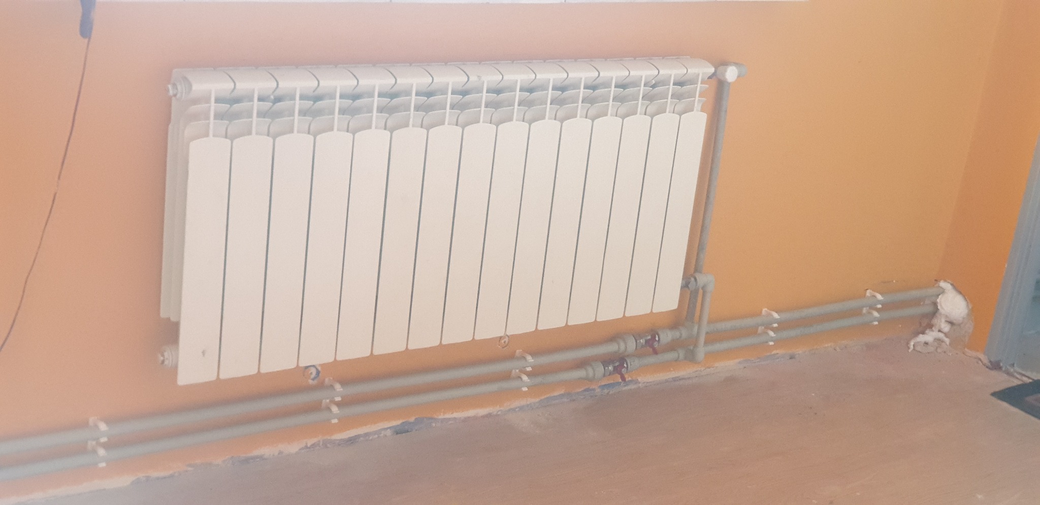 Biały grzejnik panelowy zamontowany na pomarańczowej ścianie, widoczne szare rury instalacji centralnego ogrzewania z zaworami, ściana i podłoga wymagają wykończenia.