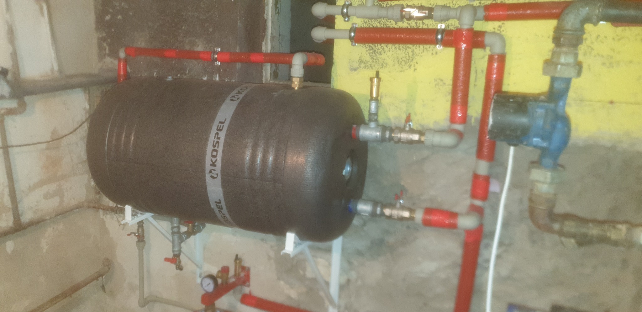 Czarny zbiornik akumulacyjny Kospel z podłączonymi rurami w kolorze czerwonym i szarym, zamontowany na białym wsporniku, widoczne zawory i pompa obiegowa na tle surowych ścian i żółtej ściany.