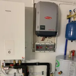 Instalacja grzewcza w kotłowni: biały kocioł gazowy Panasonic Aquarea, szary falownik Fronius Symo, zbiornik wyrównawczy i zasobnik ciepłej wody Joule, połączone rurami.