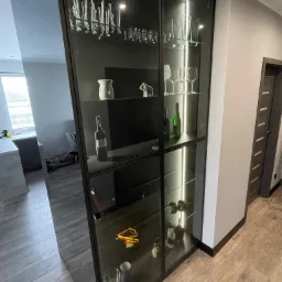 Nowoczesna, wysoka witryna z czarnymi ramami i szklanymi półkami, eksponująca kolekcję szkła, butelki wina i dekoracyjne przedmioty w oświetlonym wnętrzu.