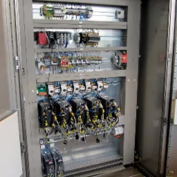 Otwarta szafa sterownicza z zamontowanymi elementami automatyki, widoczne liczne przewody, bezpieczniki i sterowniki, ułożone w uporządkowany sposób.