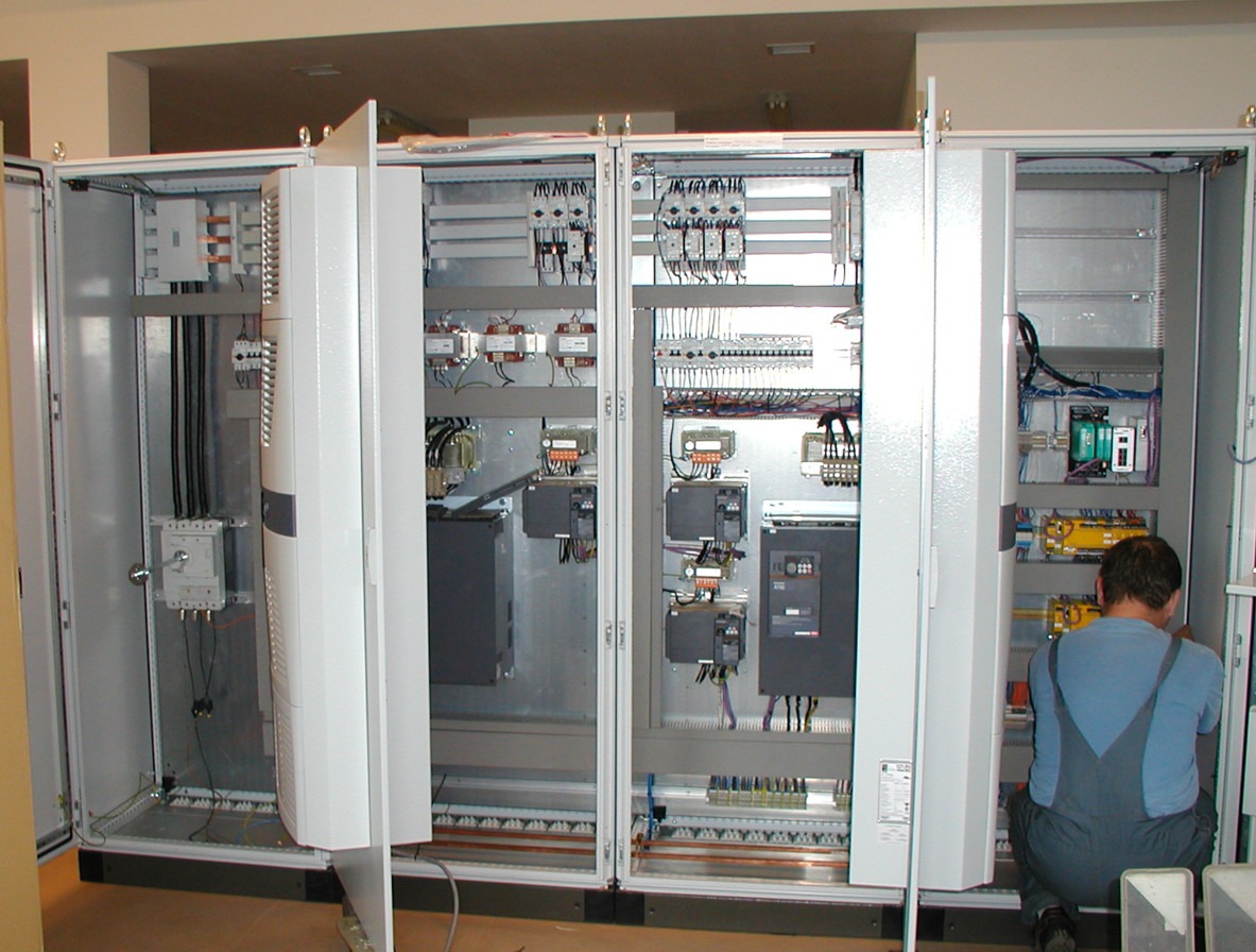 Elektryk w kombinezonie montuje lub serwisuje rozdzielnicę elektryczną w dużej metalowej szafie sterowniczej z otwartymi drzwiami, widoczne liczne przewody i zabezpieczenia.
