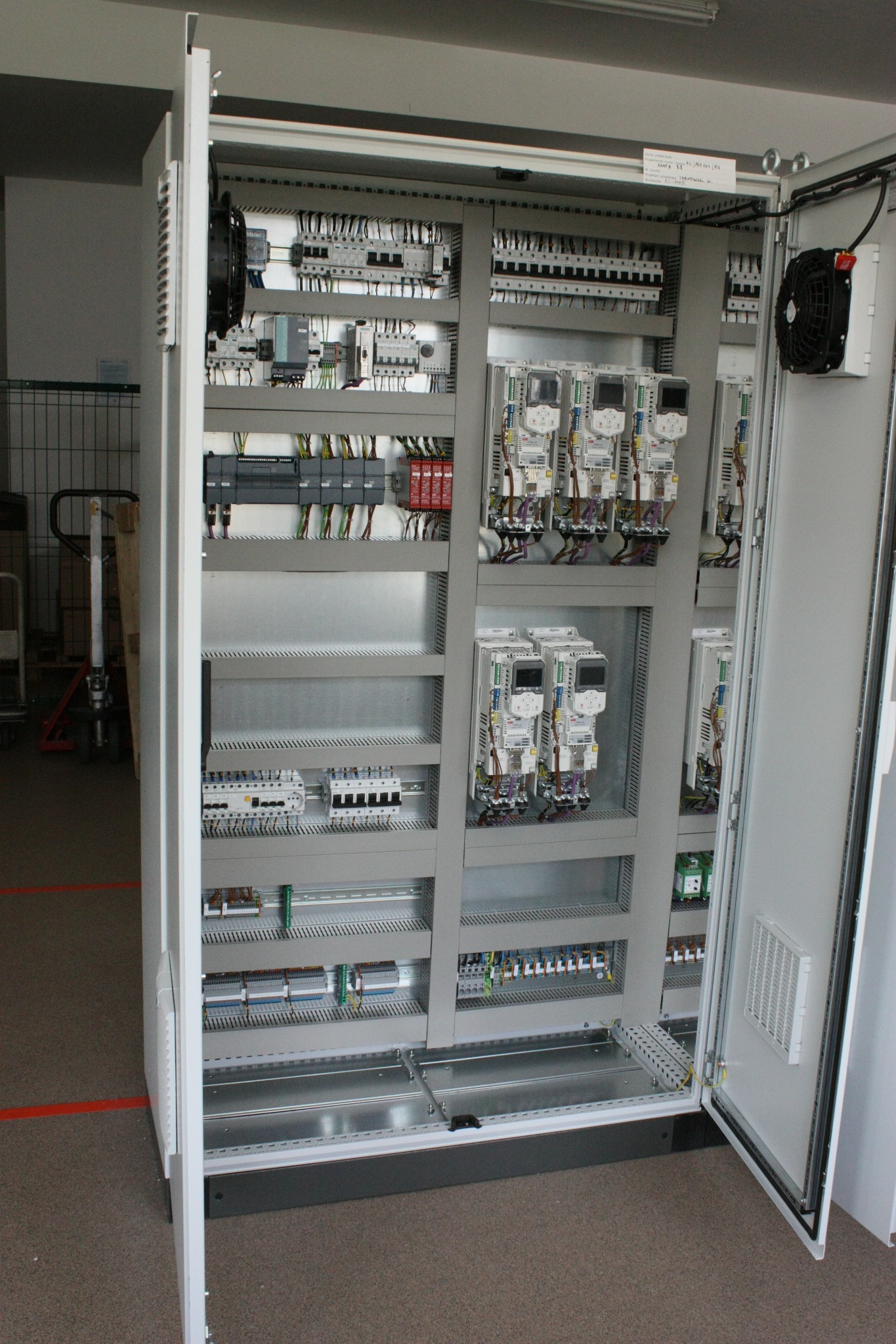 Otwarta szafa sterownicza z widocznymi modułami PLC, zasilaczami, przekaźnikami i okablowaniem, wentylator na drzwiach, widok z boku.