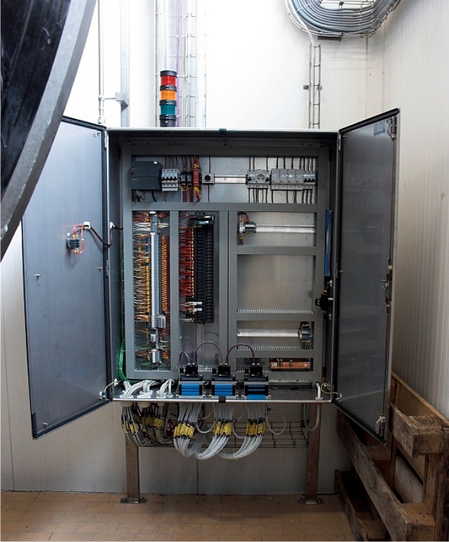 Otwarta szafa sterownicza z widocznymi przewodami, bezpiecznikami i elementami automatyki, zamontowana na metalowym stelażu w pomieszczeniu przemysłowym, z widoczną sygnalizacją świetlną na górze.