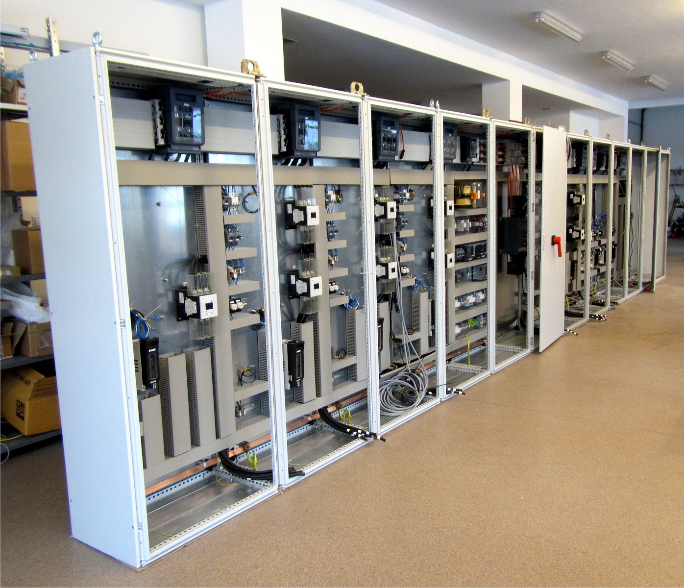 Szereg rozdzielnic elektrycznych w białych obudowach z widocznymi elementami sterowania i okablowaniem, ustawionych w rzędzie w jasnym pomieszczeniu.