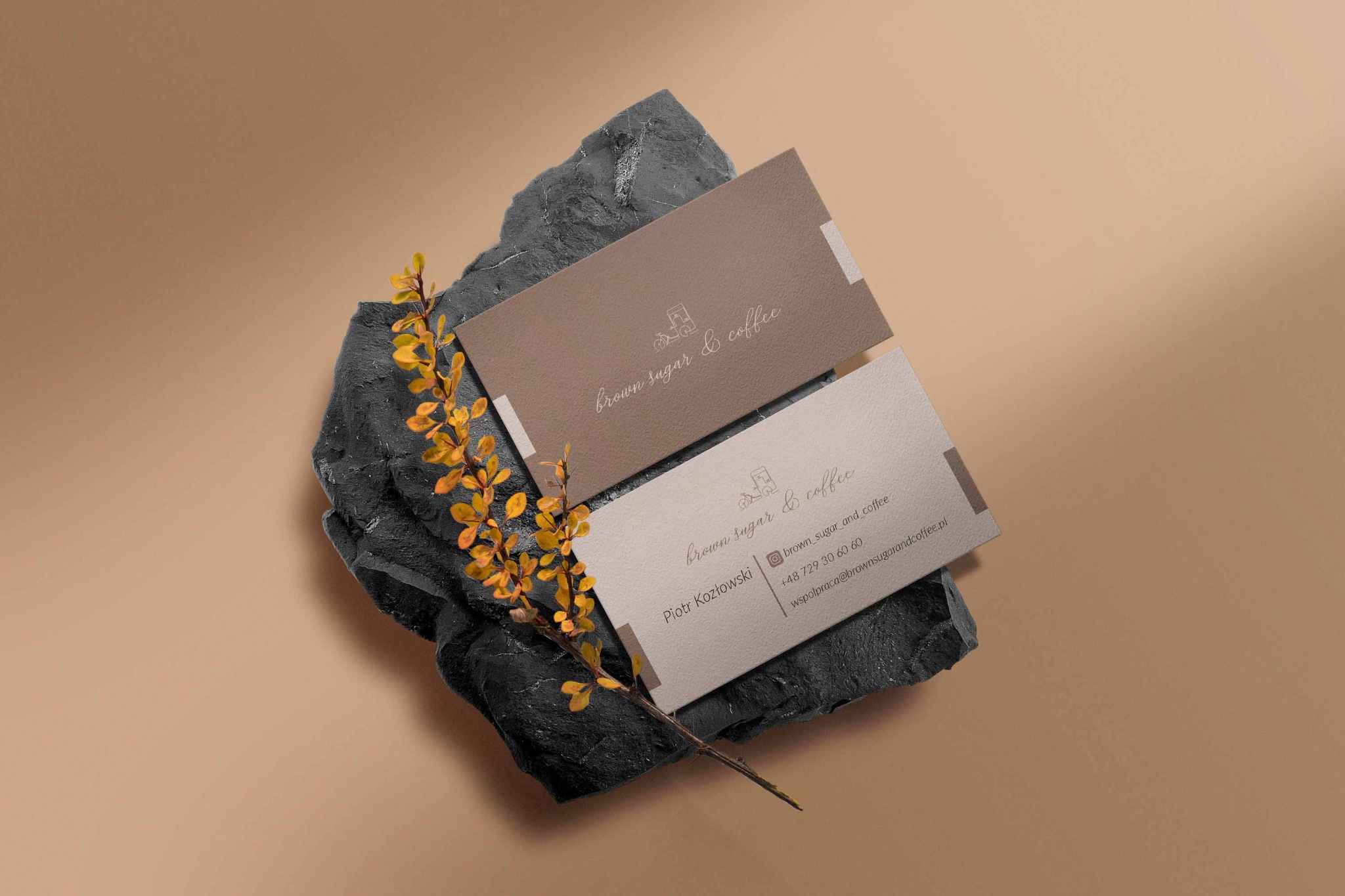 Dwie wizytówki firmy Brown Sugar & Coffee ułożone na czarnym kamieniu dekoracyjnym z gałązką z żółtymi liśćmi, na beżowym tle.