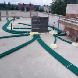 Instalacja zielonych rur wentylacyjnych na betonowym dachu budynku w trakcie budowy, zabezpieczonych pianką montażową, z widocznymi elementami konstrukcyjnymi i ceglanymi ścianami.