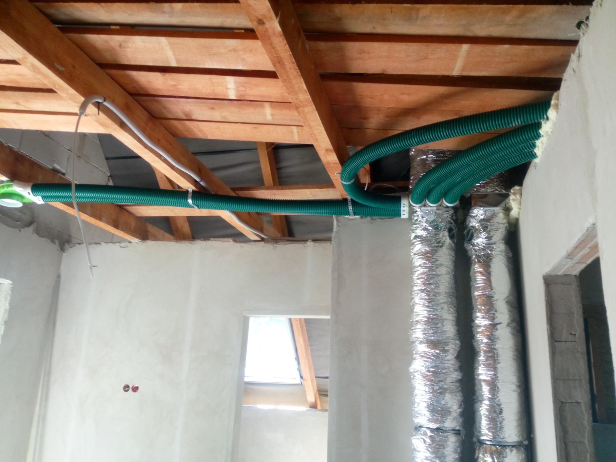 Zielone, karbowane rury wentylacyjne pod drewnianym stropem, połączone z izolowanymi, srebrnymi kanałami w surowym wnętrzu budynku w trakcie wykończenia.