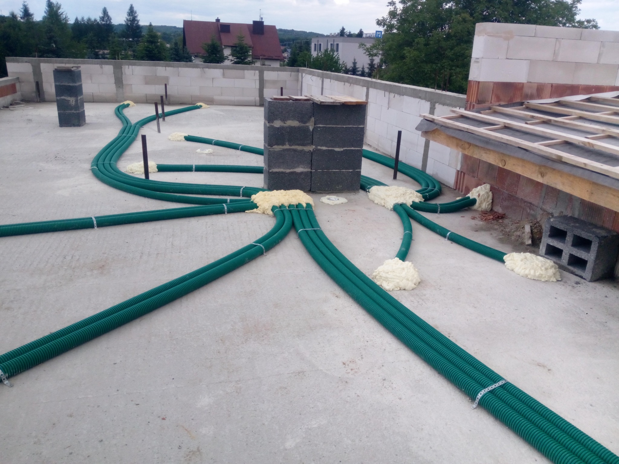 Instalacja zielonych rur wentylacyjnych na betonowym dachu budynku w trakcie budowy, zabezpieczonych pianką montażową, z widocznymi elementami konstrukcyjnymi i ceglanymi ścianami.