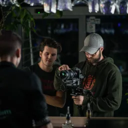 Kamerzysta z brodą i okularami, w zielonej bluzie z kapturem i szarej czapce, obsługuje profesjonalną kamerę Sony na stabilizatorze, w tle drugi mężczyzna i bar z wiszącymi kieliszkami