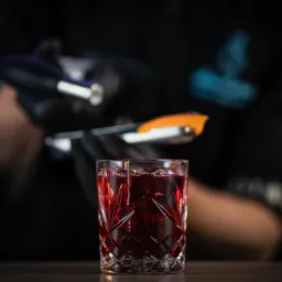 Barman w czarnych rękawicach przygotowuje drink Old Fashioned z lodem i skórką pomarańczy, widoczne szczypce barmańskie.