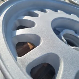 Szczegółowe ujęcie aluminiowej felgi samochodowej po obróbce, pokrytej jednolitą, matową, srebrną powłoką. Widoczne otwory wentylacyjne i delikatna faktura powierzchni.