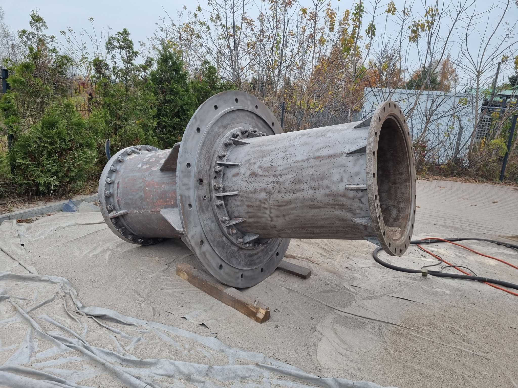 Duży, metalowy element rurowy z kołnierzami, prawdopodobnie po piaskowaniu, leżący na folii ochronnej na zewnątrz.
