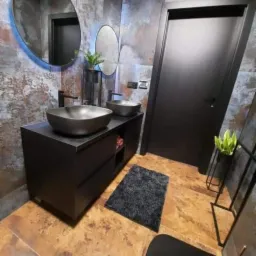 Nowoczesna łazienka z dwoma czarnymi umywalkami nablatowymi, owalnym lustrem z podświetleniem LED, czarnymi drzwiami i dekoracyjną ścianą w odcieniach brązu i szarości.