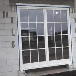 Białe, dwuskrzydłowe okno z białymi szprosami, zamontowane w surowym murze z betonu komórkowego, z widoczną pianką montażową i niebieską taśmą zabezpieczającą, odblask budynku w szybach.