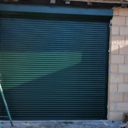 Ciemnozielona, metalowa osłona okienna zamontowana w surowym otworze budynku z widocznymi pustakami i wspornikami montażowymi.