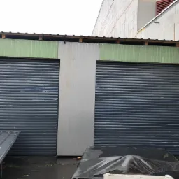 Dwie zamknięte, szare bramy rolowane w hali przemysłowej, widoczna drabina oparta o ścianę, elementy konstrukcyjne dachu i fragmenty materiałów budowlanych.