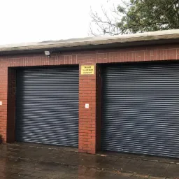 Trzy bramy garażowe rolowane z ceglaną elewacją, tabliczka informacyjna 'Ausfahrt Tag und Nacht freihalten!' na środkowej bramie, palety w tle po lewej stronie, łopata oparta o ścianę po prawej...