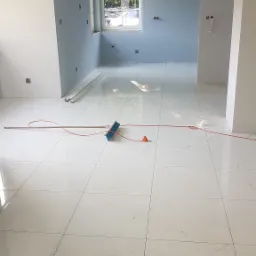 Wnętrze po remoncie z nowymi, błyszczącymi białymi płytkami podłogowymi z szarymi żyłkami, świeżo pomalowanymi na jasnoniebiesko ścianami, oknem z widokiem na zieleń oraz leżącymi na podłodze...