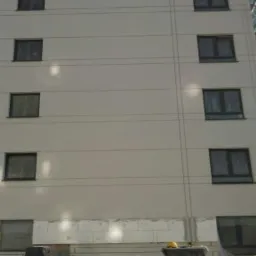 Wielopiętrowy budynek z widocznym ociepleniem fundamentów styropianem i przygotowanymi materiałami budowlanymi na pierwszym planie. Ściana budynku pomalowana na jasny kolor, widoczne okna.