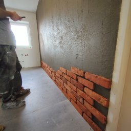 OPTIMUM kompleksowe wykończenia wnętrz - Montaż dekoracyjnej ściany z cegieł na zaprawie, widoczny fragment ściany z nałożoną szarą zaprawą i częściowo ułożonymi cegłami, pracownik w tle z telefonem.