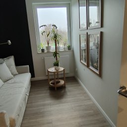 OPTIMUM kompleksowe wykończenia wnętrz - Jasne panele podłogowe imitujące drewno w pokoju z szarą ścianą i czarnym akcentem, sofa z jasną narzutą, okno z roślinami i okrągły stolik, na ścianie wiszą cztery obrazy w ramkach.