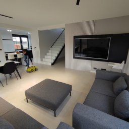 OPTIMUM kompleksowe wykończenia wnętrz - Wnętrze salonu z szarą sofą modułową i pufą, pracownik z torbą narzędziową w tle, białe schody z czarną balustradą w głębi, nowoczesne oświetlenie sufitowe.