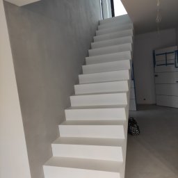 OPTIMUM kompleksowe wykończenia wnętrz - Białe, minimalistyczne schody w nowoczesnym wnętrzu, ściana wykończona gładzią w odcieniach szarości, zabezpieczone drzwi w tle.