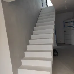 Białe, minimalistyczne schody w nowoczesnym wnętrzu, ściana wykończona gładzią w odcieniach szarości, zabezpieczone drzwi w tle.