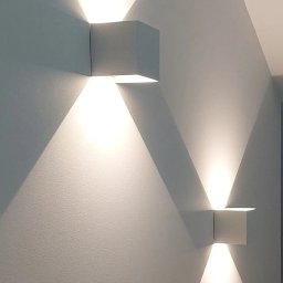 OPTIMUM kompleksowe wykończenia wnętrz - Dwa kwadratowe, białe kinkiety LED na pomalowanej na biało ścianie, emitujące trójkątne strumienie światła skierowane w górę i w dół.