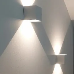 Dwa kwadratowe, białe kinkiety LED na pomalowanej na biało ścianie, emitujące trójkątne strumienie światła skierowane w górę i w dół.
