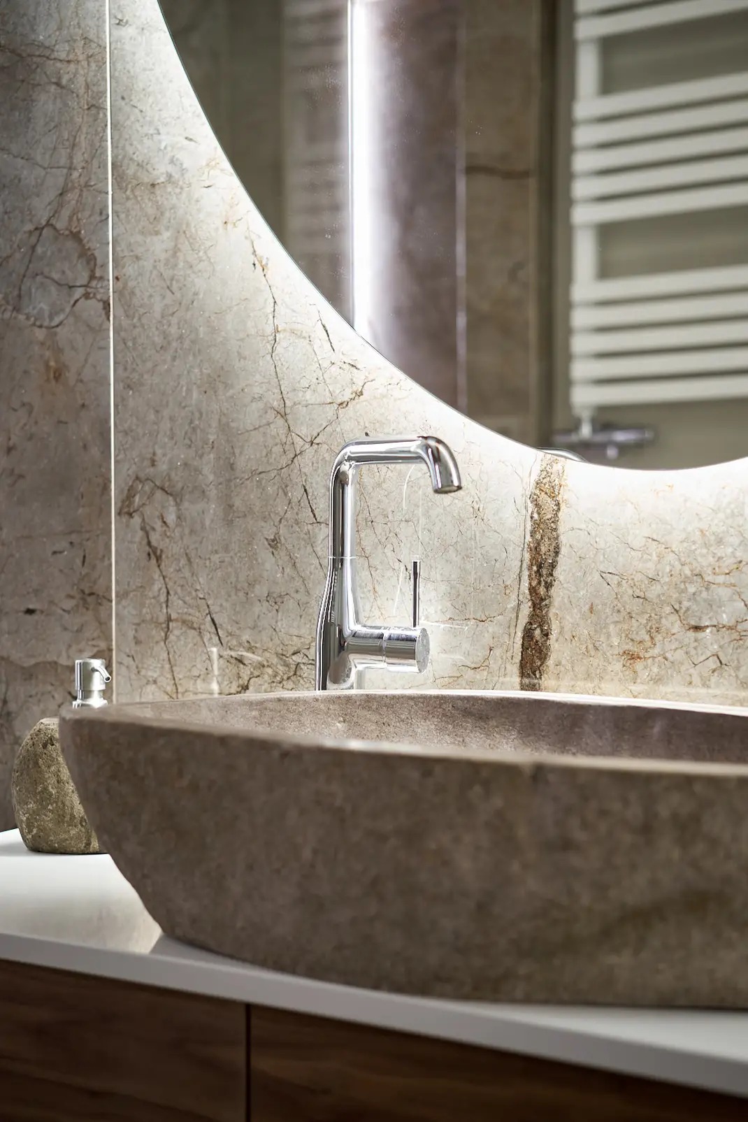 Nowoczesna łazienka z umywalką z naturalnego kamienia, chromowaną baterią i lustrem w marmurowej ramie, uzupełniona o dozownik mydła z kamienia i grzejnik drabinkowy w tle.