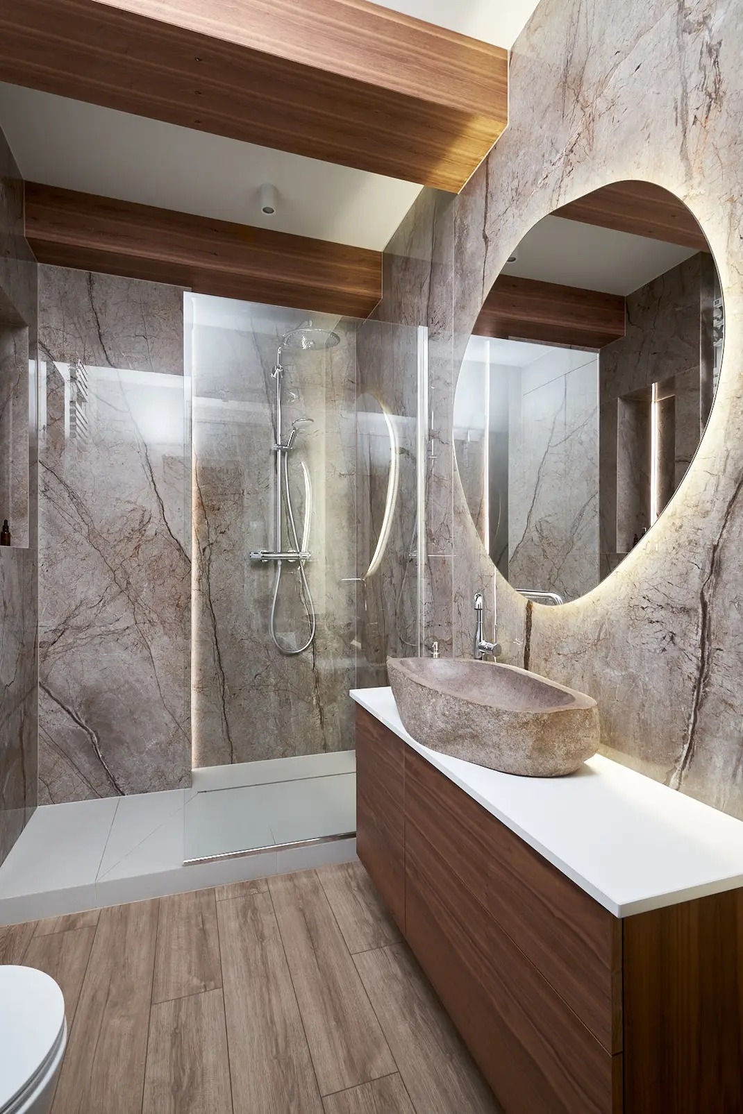 Nowoczesna łazienka z marmurowymi ścianami, kabiną prysznicową ze szklanymi drzwiami, umywalką z kamienia na drewnianej szafce z białym blatem i okrągłym lustrem z podświetleniem LED.