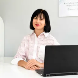 Uśmiechnięta kobieta w koszuli w paski siedzi za biurkiem z laptopem, w tle certyfikat i informacja o kupnie/sprzedaży nieruchomości.