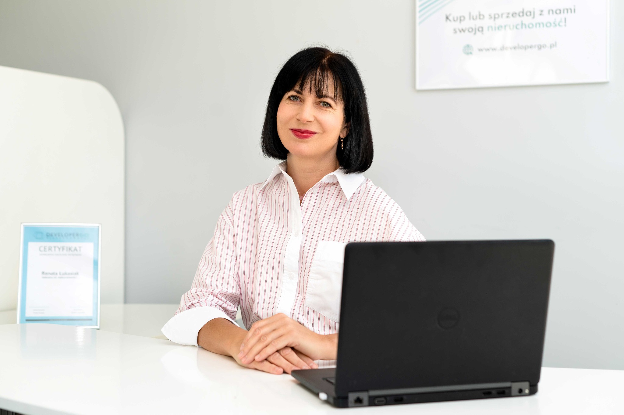 Uśmiechnięta kobieta w koszuli w paski siedzi za biurkiem z laptopem, w tle certyfikat i informacja o kupnie/sprzedaży nieruchomości.