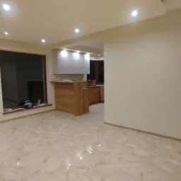 Jasne wnętrze po remoncie: marmurowe płytki podłogowe, świeżo pomalowane ściany w odcieniach beżu, widoczna kuchnia z drewnianymi szafkami i barek, okno z narzędziami na parapecie, sufit...