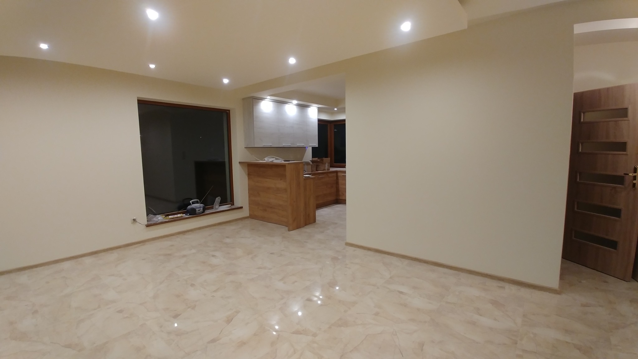 Jasne wnętrze po remoncie: marmurowe płytki podłogowe, świeżo pomalowane ściany w odcieniach beżu, widoczna kuchnia z drewnianymi szafkami i barek, okno z narzędziami na parapecie, sufit...