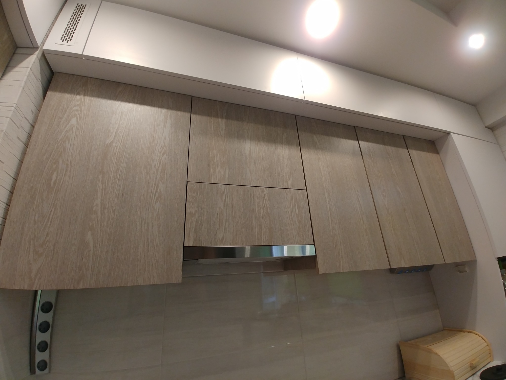 Jasne, drewnopodobne szafki kuchenne wiszące nad blatem z beżowych płytek, z widocznym okapem i listwą z gniazdkami.