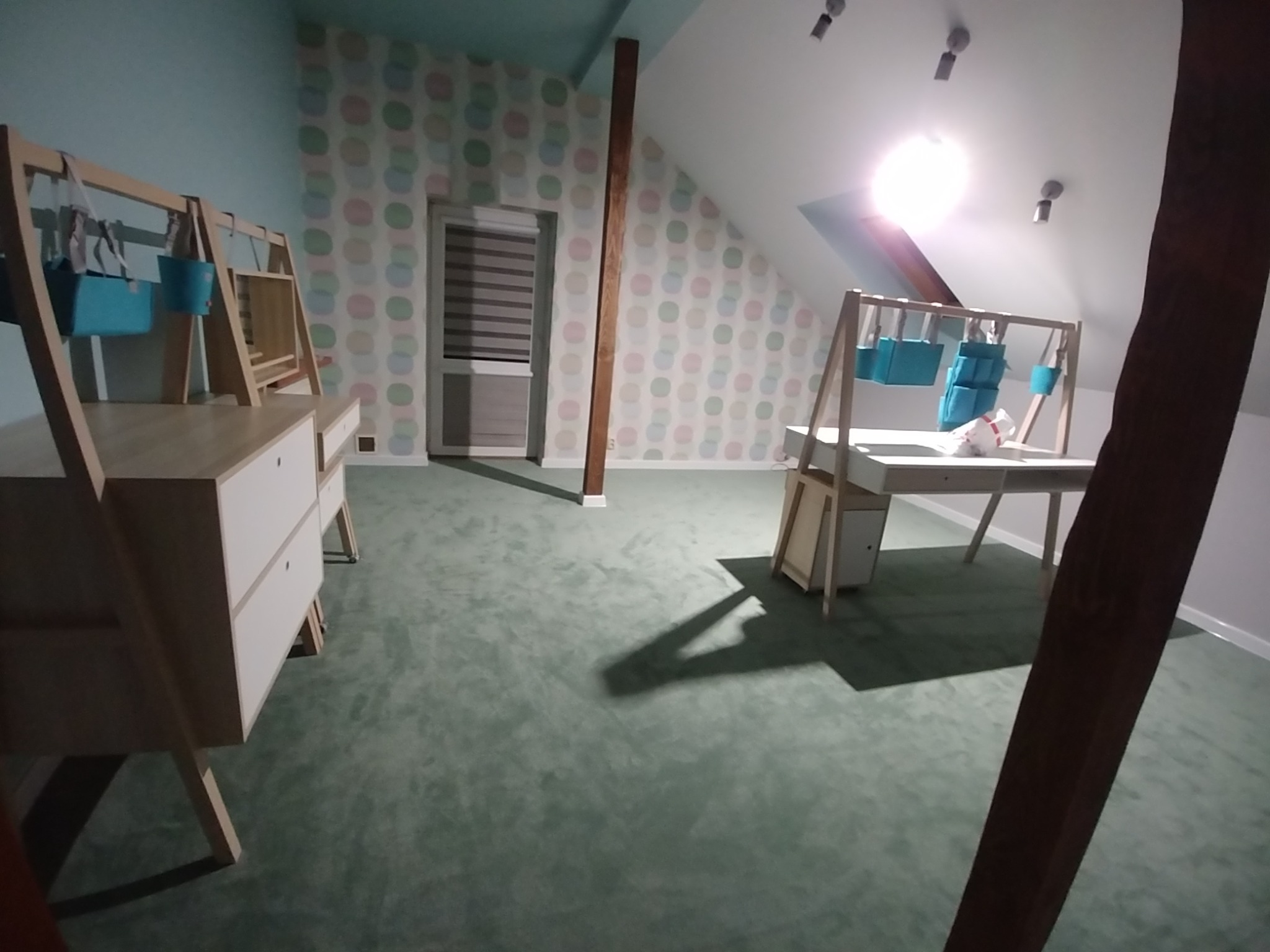 Pokój na poddaszu z meblami dziecięcymi: biurko z organizerem na zabawki, komoda z szufladami, ściany w pastelowe kropki, drewniana belka konstrukcyjna.