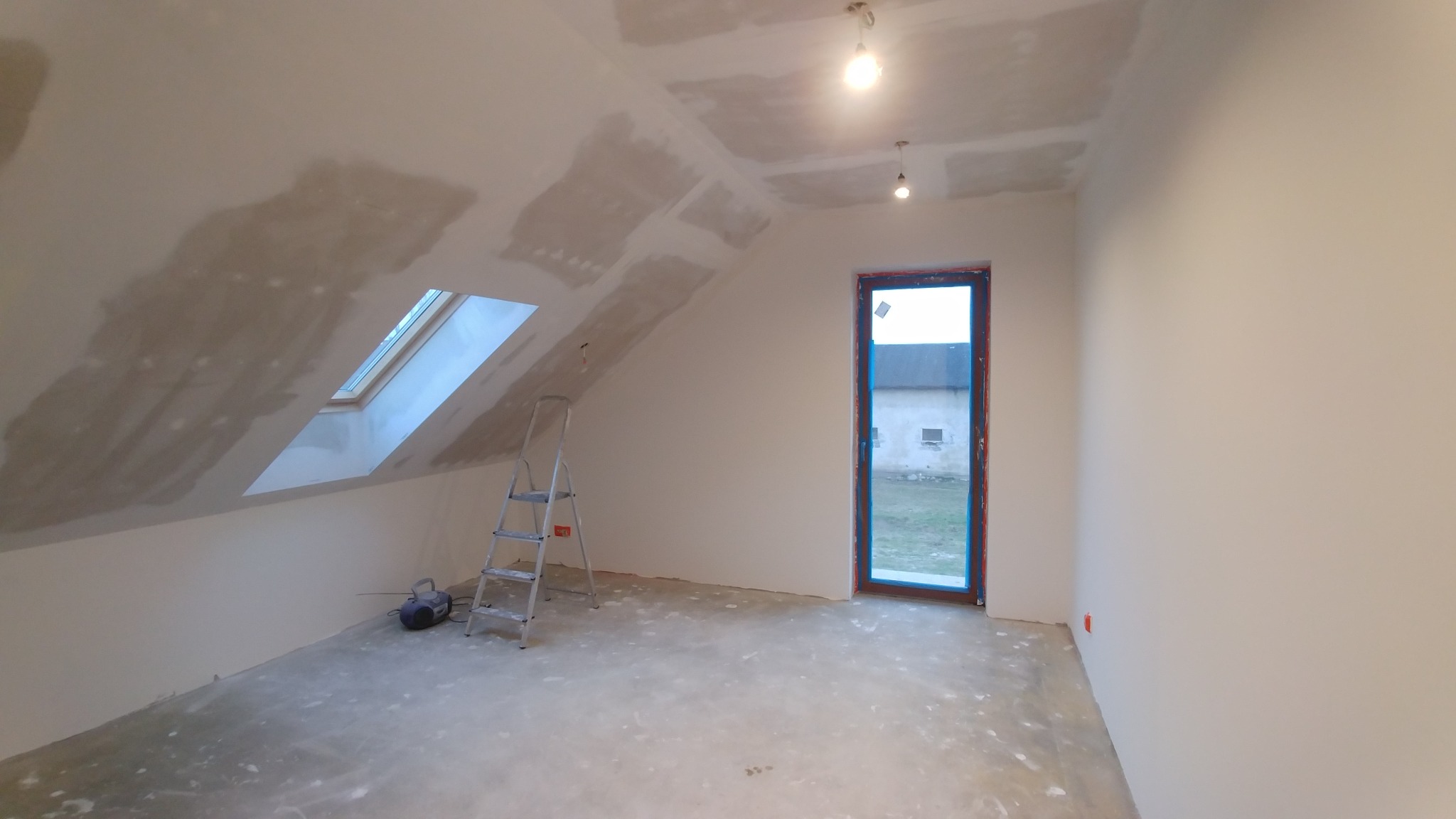 Poddasze w trakcie remontu: szpachlowane ściany i sufit ze świetlikiem, drabina aluminiowa i okno z niebieską folią ochronną.