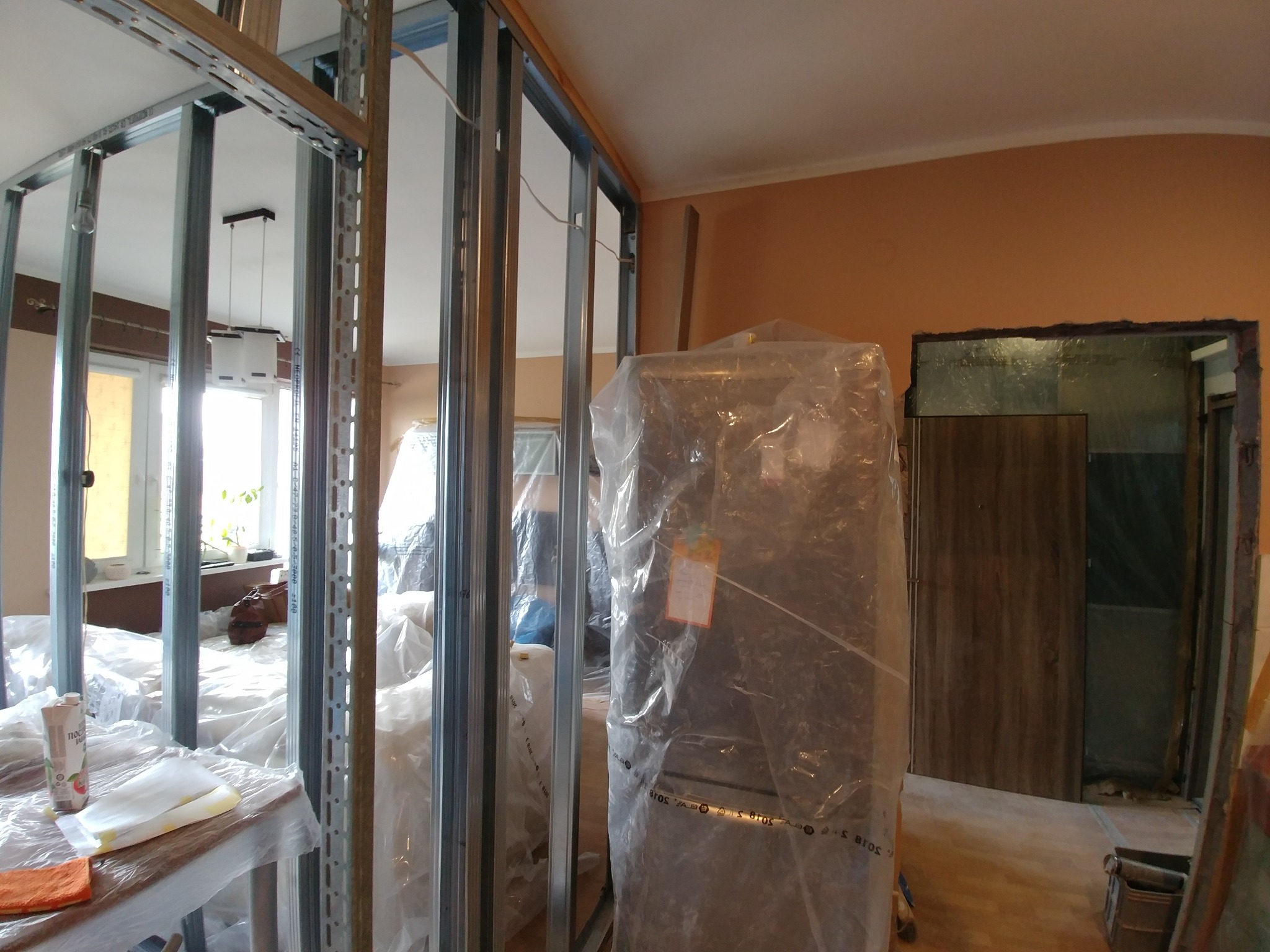 Wnętrze w trakcie remontu: metalowa konstrukcja ścianki działowej, zabezpieczona folią lodówka, widoczne elementy wyposażenia przykryte folią malarską.