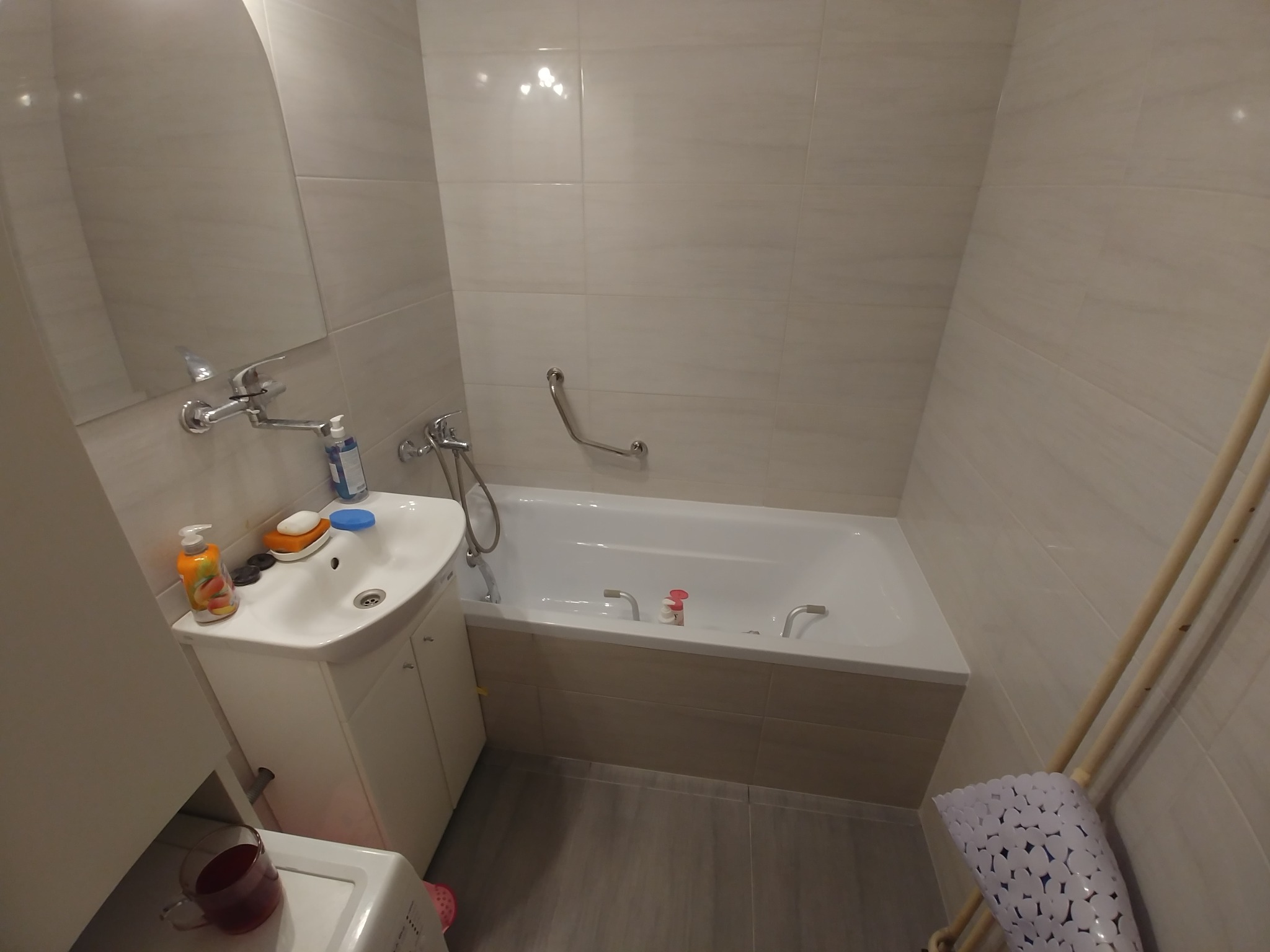 Wykończona łazienka z białymi płytkami, wanną z obudową z płytek imitujących drewno, umywalką z szafką i lustrem, wyposażona w uchwyt dla niepełnosprawnych oraz kosz na pranie.