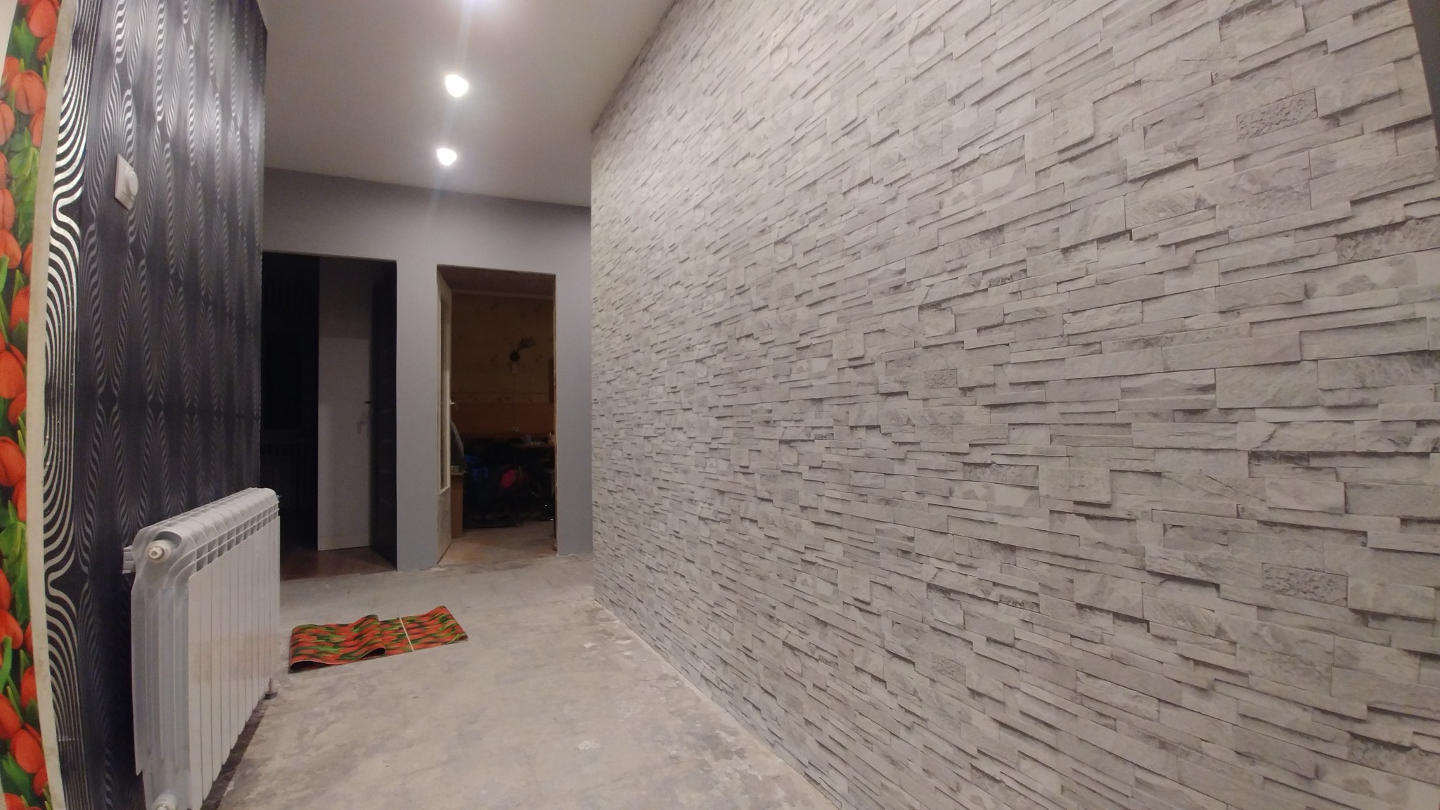Korytarz z wykończoną ścianą z dekoracyjnej szarej cegły, widoczny grzejnik, wzorzysta tapeta i dywanik na podłodze.