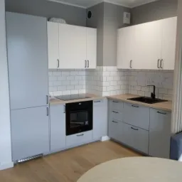 Nowoczesna kuchnia w odcieniach szarości i bieli, z drewnianym blatem i podłogą, oświetlona reflektorami sufitowymi, z widocznymi planami technicznymi na ścianie.