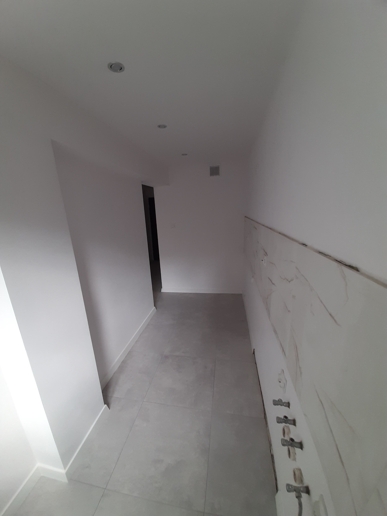 Korytarz w trakcie prac wykończeniowych: położone szare płytki podłogowe, ściany pomalowane na biało, z widocznym fragmentem ściany z imitacją marmuru i wystającymi rurami instalacji wodnej przed...