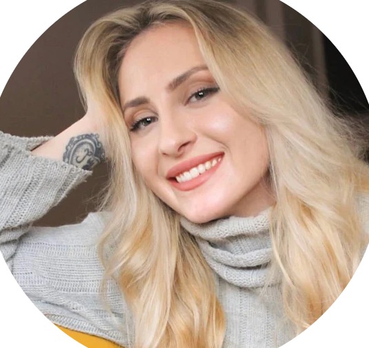 Portret uśmiechniętej blondynki w szarym swetrze z golfem, z tatuażem na nadgarstku, spoglądającej w obiektyw.